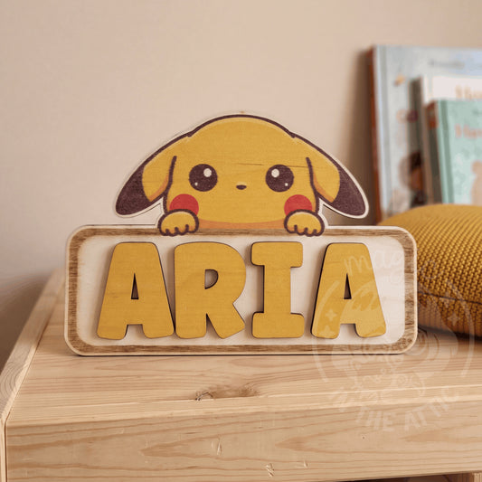 Placa de madera personalizada con Pikachu asomando por encima del nombre Aria en letras amarillas en relieve, apoyada en una estantería de madera junto a un cojín mostaza