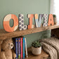 Letras de madera Bluey OLIVIA apoyadas en estantería con libros y peluches al fondo
