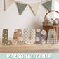 Letras de madera personalizadas con estampado de ovejitas y flores formando el nombre Laura, sobre balda de madera clara en habitación infantil