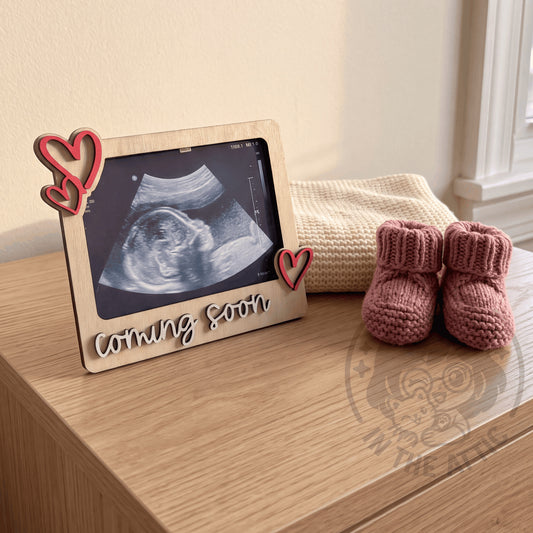 Marco de madera personalizado Coming Soon con corazones rosa y ecografía sobre cómoda en habitación de bebé en preparación