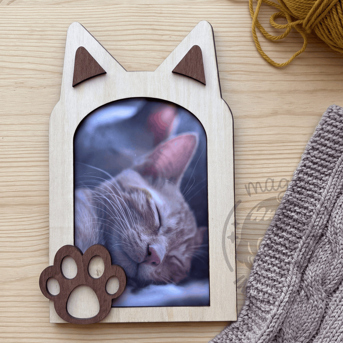 Marco de fotos con forma de gato en madera vista cenital sobre mesa de madera clara junto a manta y ovillo de lana