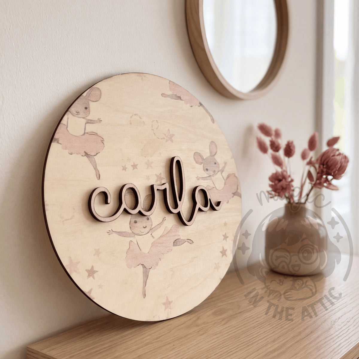 Placa de madera redonda con el nombre Carla y estampado de ratoncitas bailarinas, apoyada en una cómoda junto a un espejo redondo y un jarrón con flores secas rosas