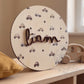 Placa de madera redonda con el nombre Liam y estampado de cochecitos, apoyada en una estantería de madera clara junto a un cojín mostaza y un juguete de madera