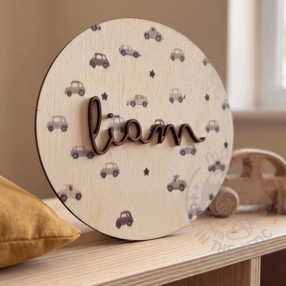 Placa de madera redonda con el nombre Liam y estampado de cochecitos, apoyada en una estantería de madera clara junto a un cojín mostaza y un juguete de madera