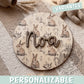 Placa de madera redonda personalizada con el nombre Noa en letras de madera oscura en relieve, sobre un fondo impreso de conejitos de acuarela entre hierbas, encima de una manta de punto junto a espigas secas y un peluche