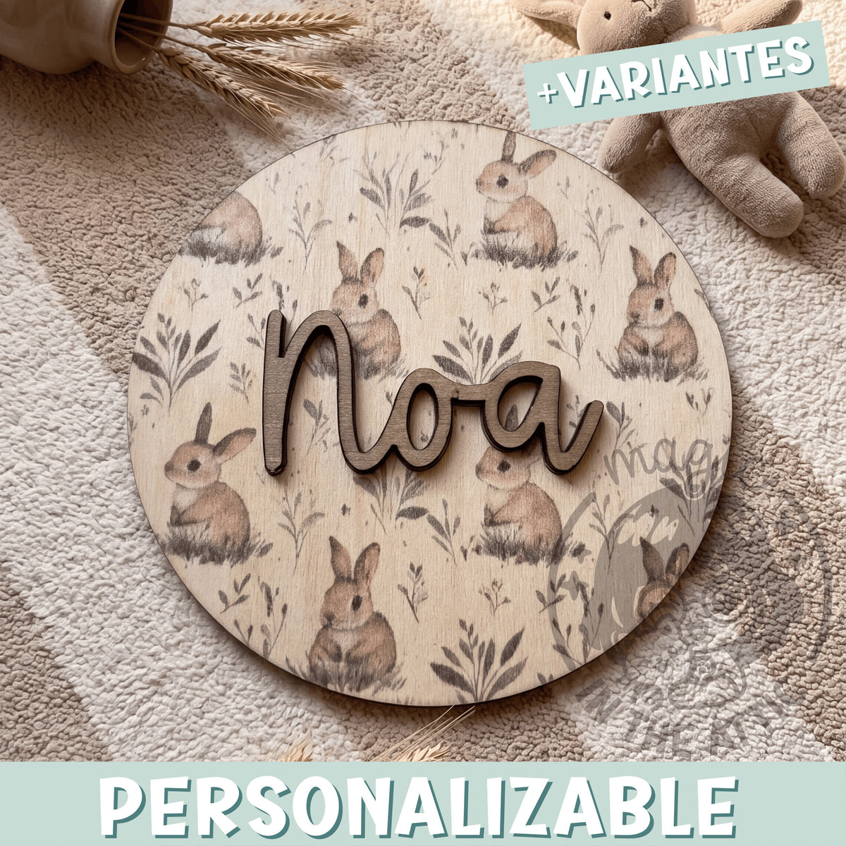 Placa de madera redonda personalizada con el nombre Noa en letras de madera oscura en relieve, sobre un fondo impreso de conejitos de acuarela entre hierbas, encima de una manta de punto junto a espigas secas y un peluche
