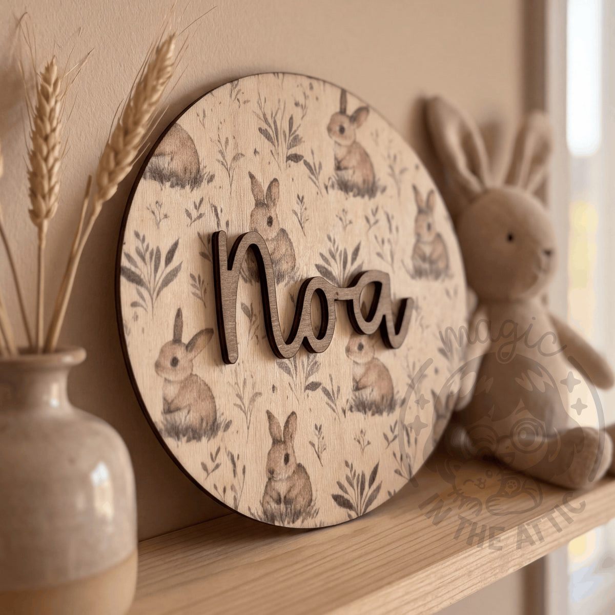 Placa de madera redonda con el nombre Noa y estampado de conejitos, apoyada en una estantería junto a un jarrón con espigas y un conejito de peluche