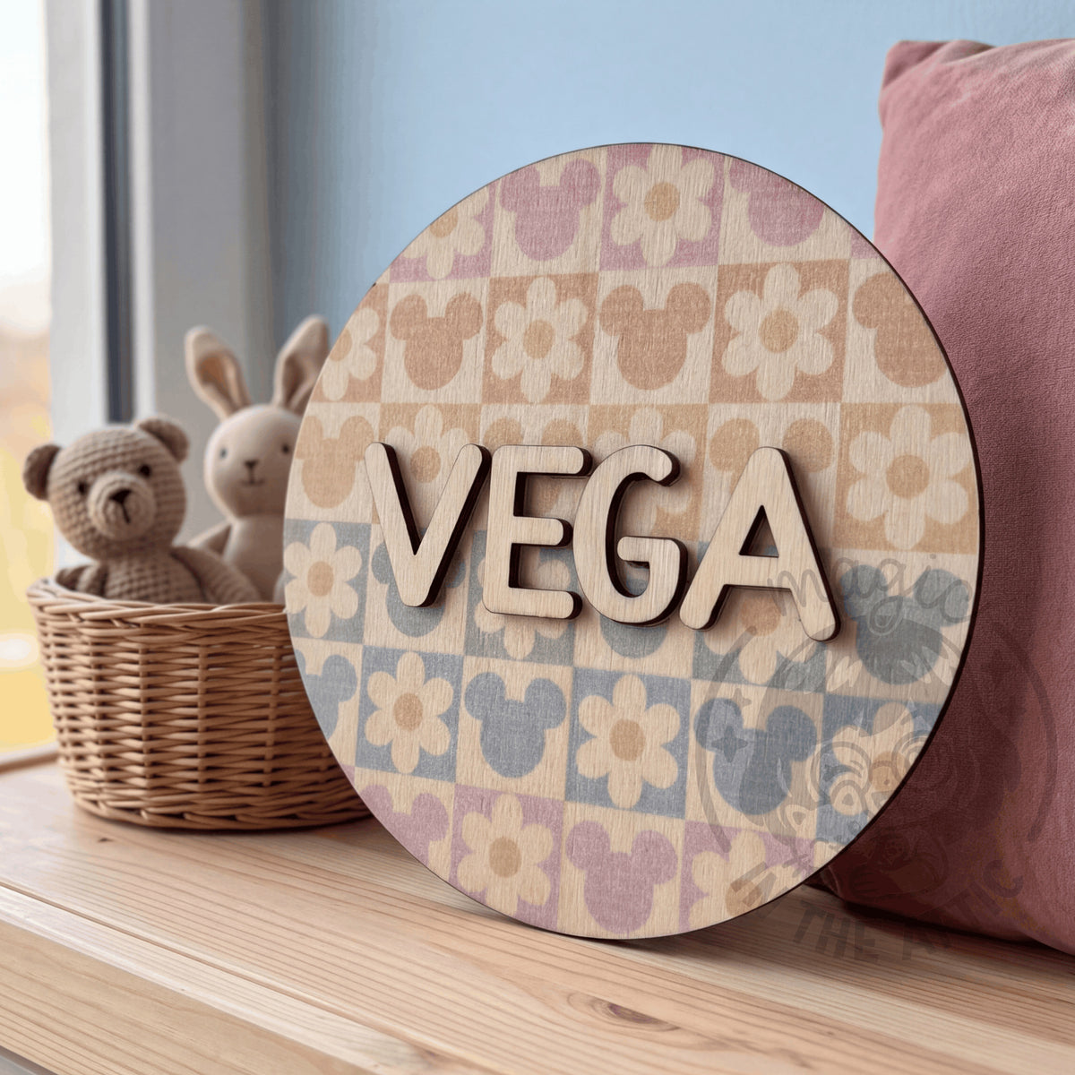 Placa de madera redonda con el nombre Vega y estampado pastel de Mickey y flores, apoyada en un banco junto a la ventana, al lado de una cesta de mimbre con dos peluches y un cojín rosa