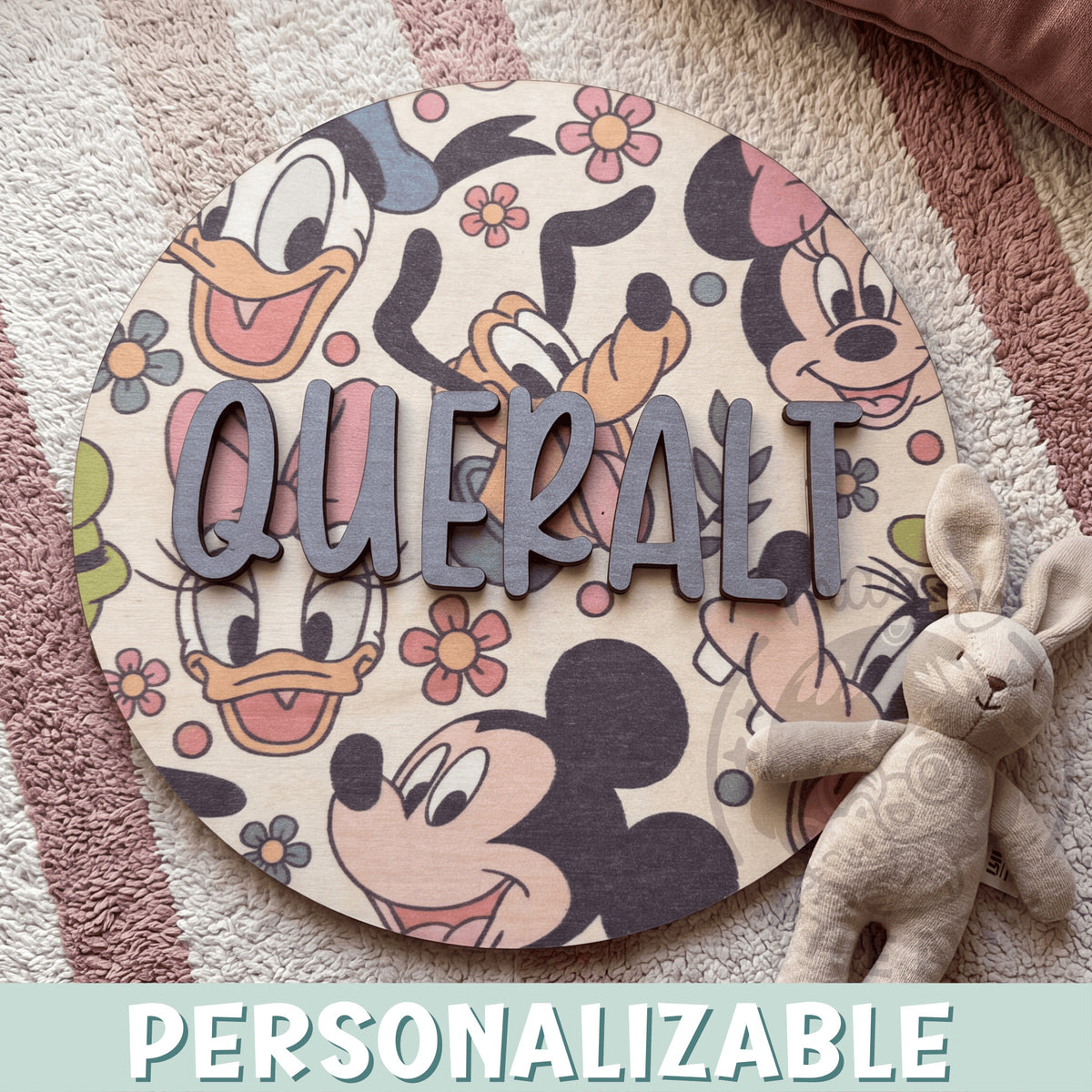 Placa de madera redonda personalizada con el nombre Queralt en letras de madera gris en relieve, sobre un fondo impreso de Mickey, Minnie, Donald, Daisy y Goofy con flores de estilo vintage, encima de una alfombra de rayas rosas junto a un conejito de peluche