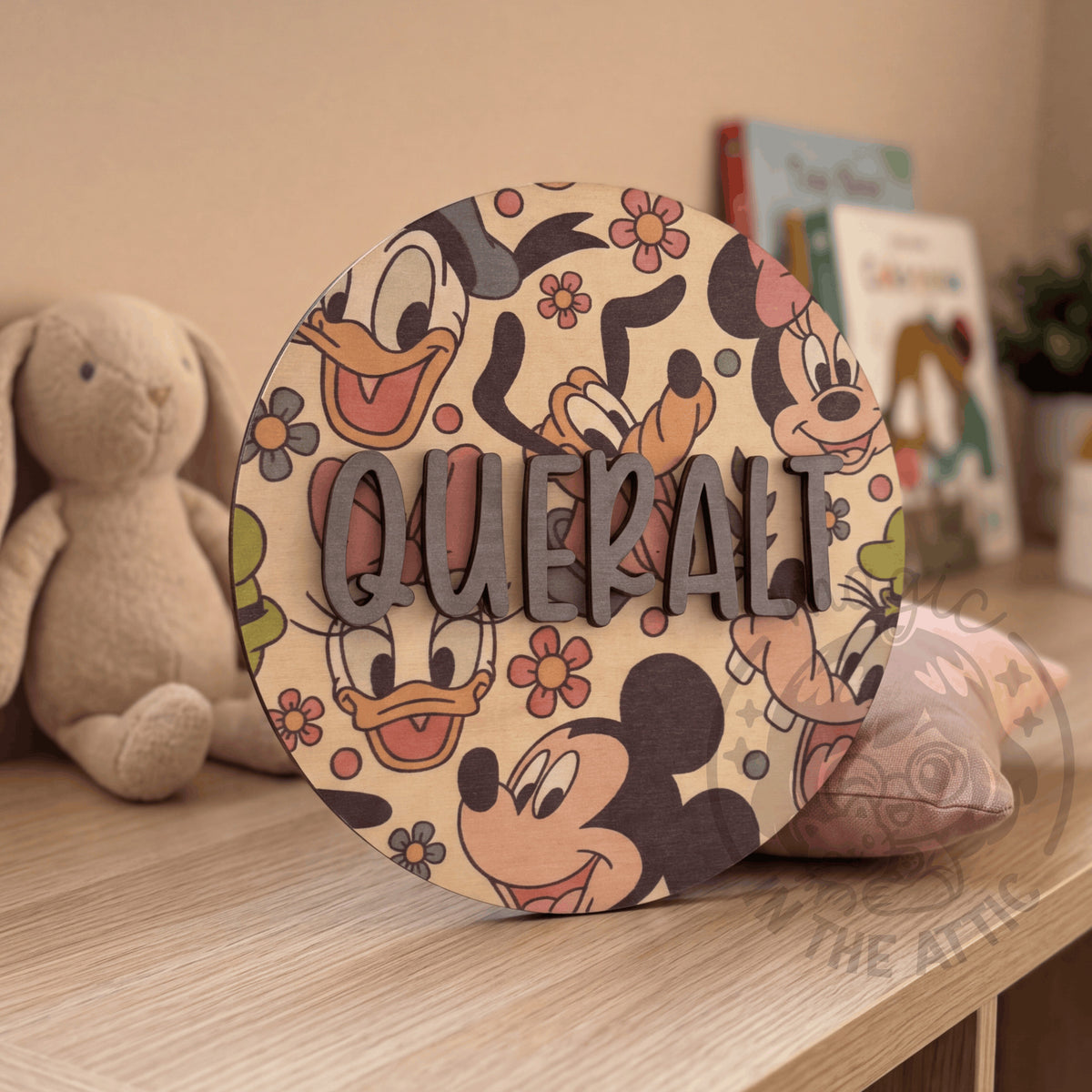 Placa de madera redonda con el nombre Queralt y estampado de Mickey y sus amigos, apoyada en una cómoda junto a un conejito de peluche y unos libros infantiles