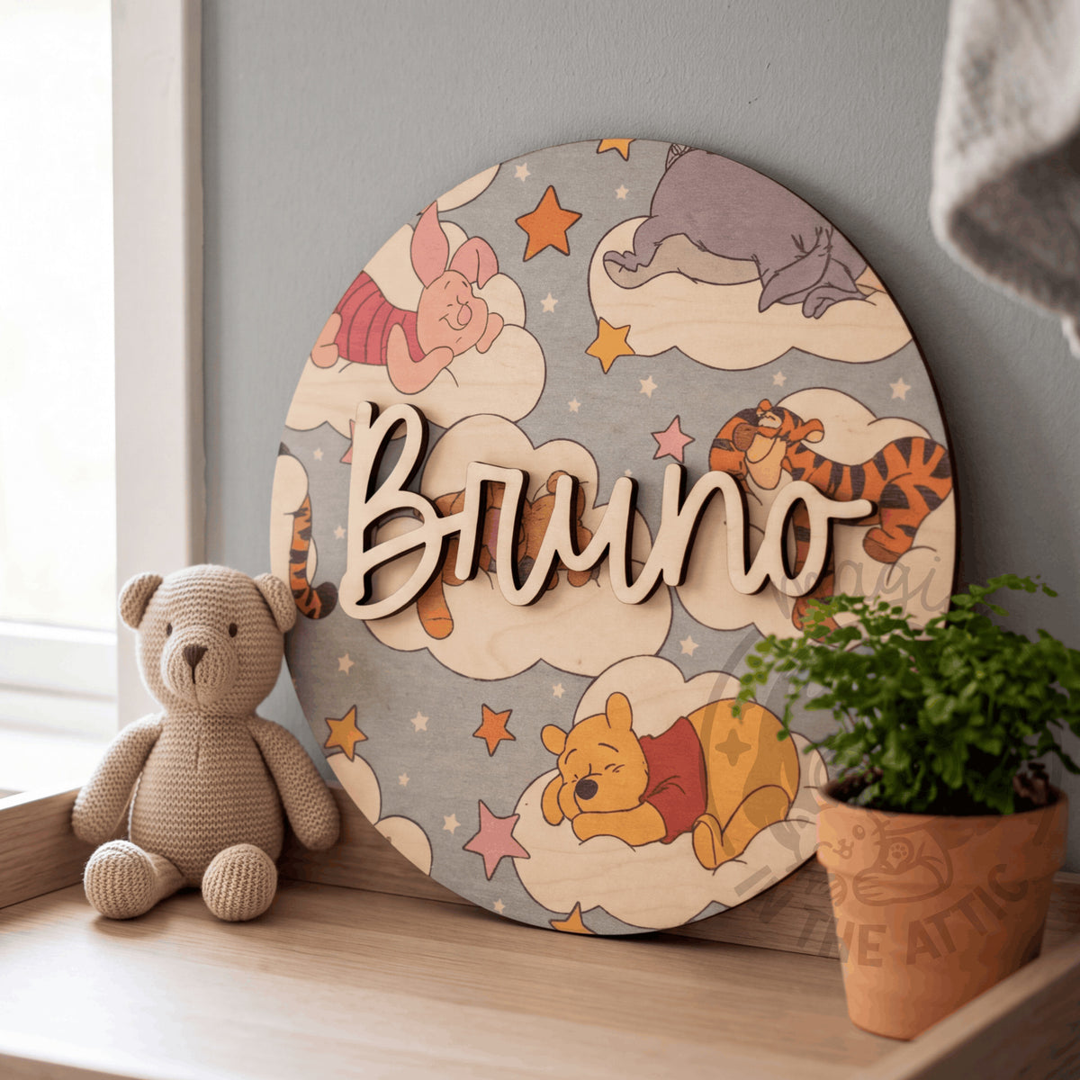 Placa de madera redonda con el nombre Bruno y estampado de Winnie the Pooh entre nubes, apoyada en una repisa junto a la ventana, entre un osito de peluche y una maceta con un helecho
