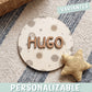 Placa de madera redonda personalizada con el nombre Hugo en letras de madera marrón en relieve, sobre un fondo impreso de planetas, soles sonrientes, arcoíris y estrellas en tonos tierra, encima de una alfombra de rayas junto a un cojín de estrella mostaza