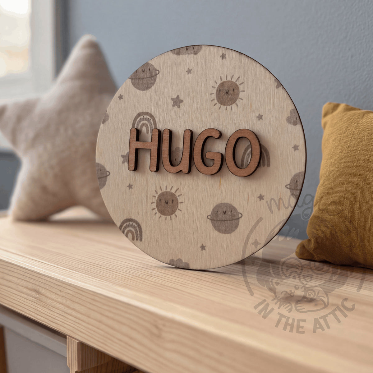 Placa de madera redonda con el nombre Hugo y estampado de galaxia en tonos suaves, apoyada en una repisa de madera entre un cojín de estrella y un cojín mostaza