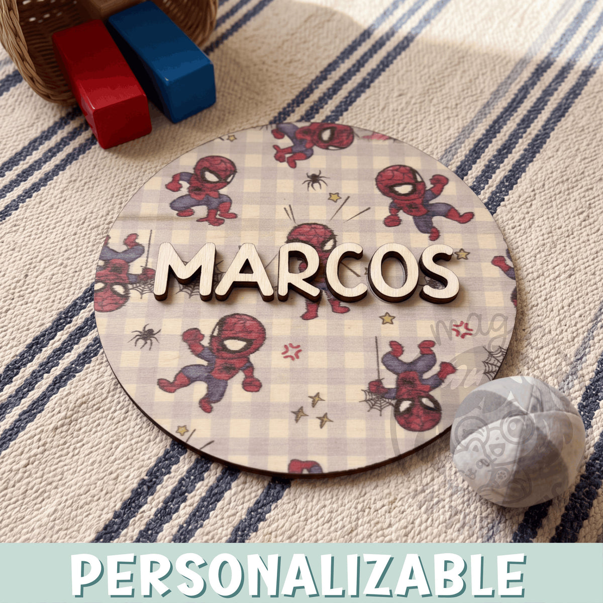 Placa de madera redonda personalizada con el nombre Marcos en letras de madera natural en relieve, sobre un fondo impreso de Spider-Man en versión infantil y arañitas sobre cuadros vichy beige, encima de una alfombra de rayas azules junto a unos bloques de madera y una pelota de tela