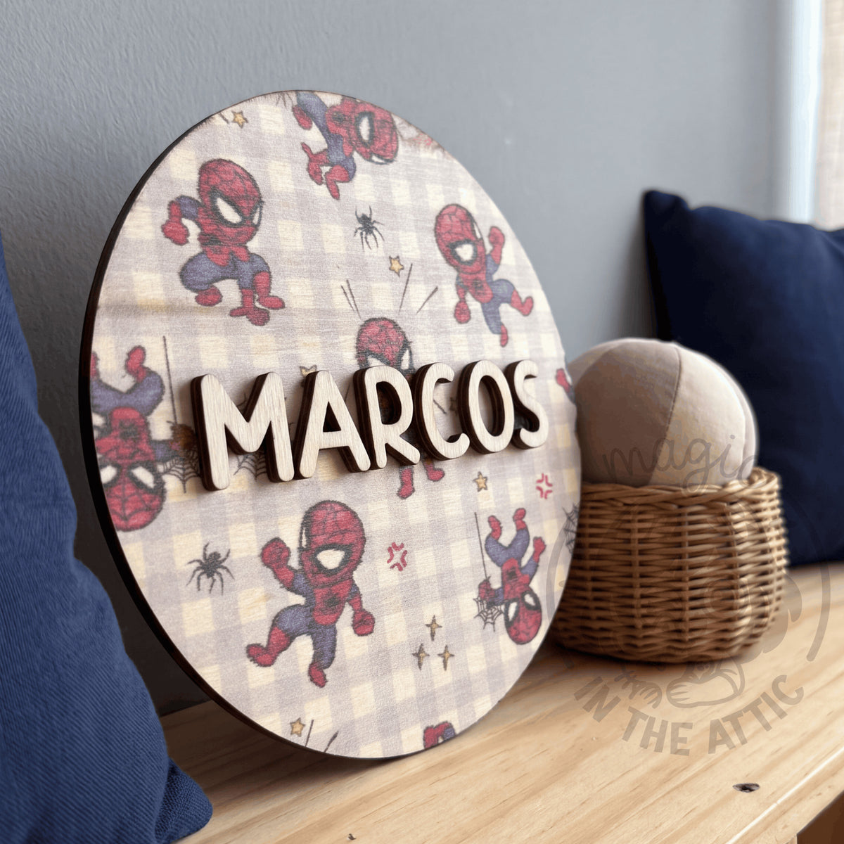 Placa de madera redonda con el nombre Marcos y estampado de Spider-Man sobre cuadros vichy, apoyada en un banco de madera junto a un cojín azul marino y una cesta de mimbre con una pelota de tela
