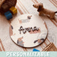 Placa de madera redonda personalizada con el nombre Anna en letras de madera oscura en relieve, sobre un fondo impreso de perritos salchicha con jersey en tonos tierra, encima de una alfombra de rayas junto a un perro de peluche y bloques de madera de colores