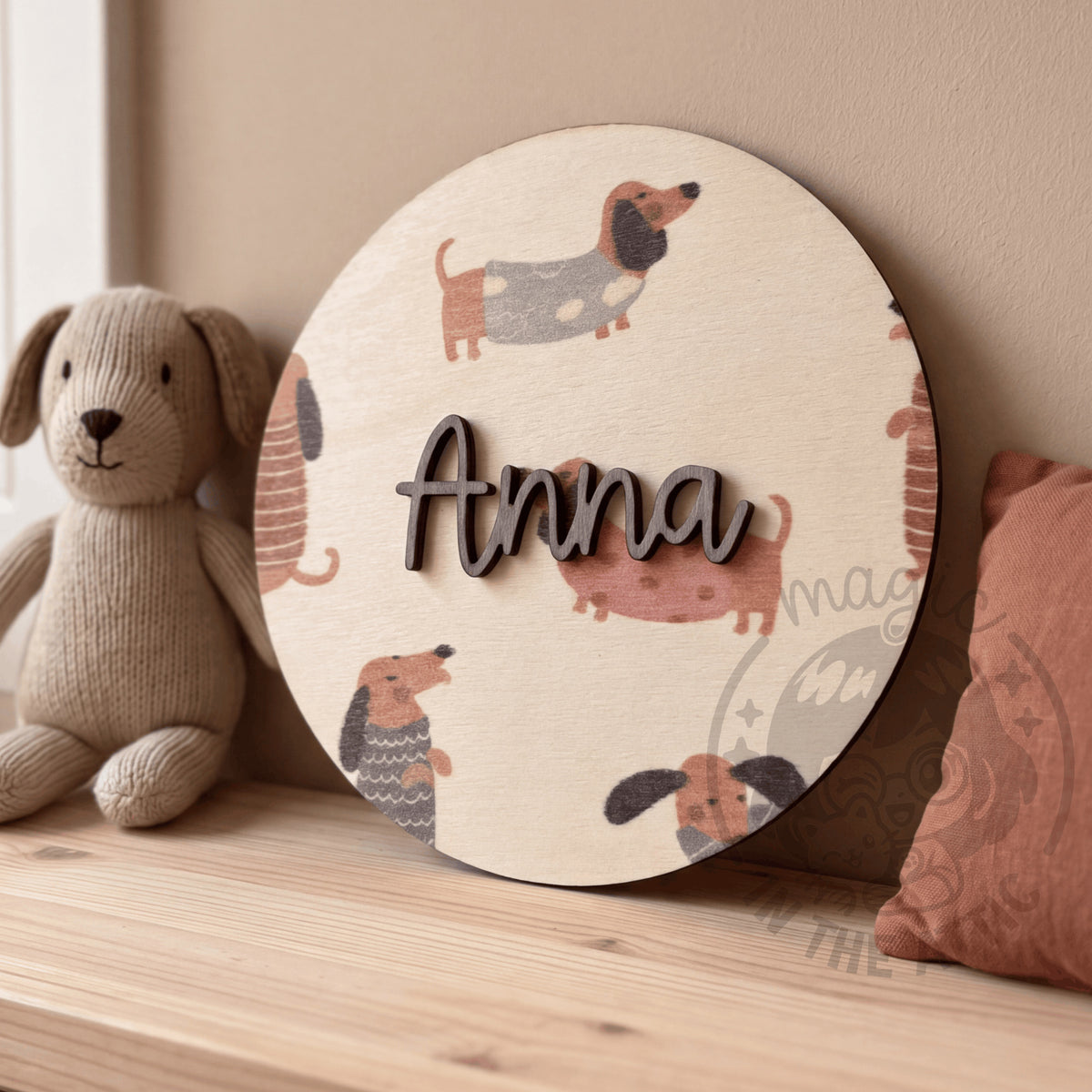 Placa de madera redonda con el nombre Anna y estampado de perritos salchicha, apoyada en un banco de madera entre un perrito de peluche de punto y un cojín color teja