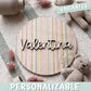 Placa de madera redonda personalizada con el nombre Valentina en letras de madera natural en relieve, sobre un fondo impreso de rayas de acuarela en tonos pastel, encima de una manta de punto junto a un osito de peluche y flores secas rosas