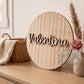 Placa de madera redonda con el nombre Valentina y estampado de rayas pastel de acuarela, apoyada en una cómoda junto a una cesta de mimbre con una muselina y un jarrón con una rosa y pampas