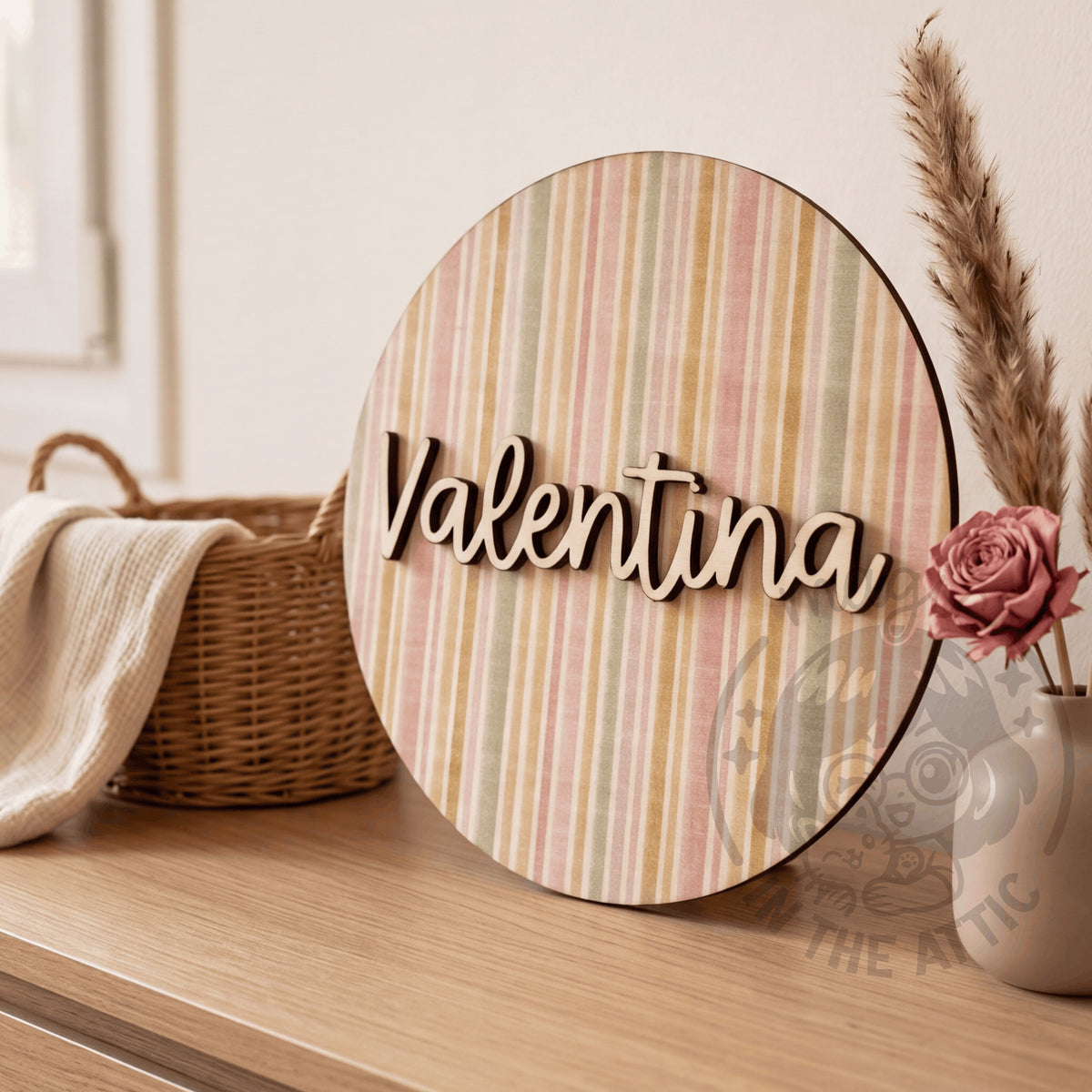 Placa de madera redonda con el nombre Valentina y estampado de rayas pastel de acuarela, apoyada en una cómoda junto a una cesta de mimbre con una muselina y un jarrón con una rosa y pampas