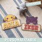 Dos placas de madera personalizadas con nombre y un Pokémon asomando por encima: Pikachu con el nombre Aria en letras amarillas y Gengar con el nombre Iker en letras rojas, sobre una alfombra de rayas azules junto a un peluche gris