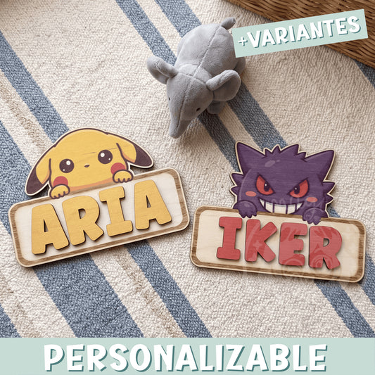Dos placas de madera personalizadas con nombre y un Pokémon asomando por encima: Pikachu con el nombre Aria en letras amarillas y Gengar con el nombre Iker en letras rojas, sobre una alfombra de rayas azules junto a un peluche gris