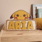 Placa de madera personalizada con Pikachu asomando por encima del nombre Aria en letras amarillas en relieve, apoyada en una estantería de madera junto a un cojín mostaza