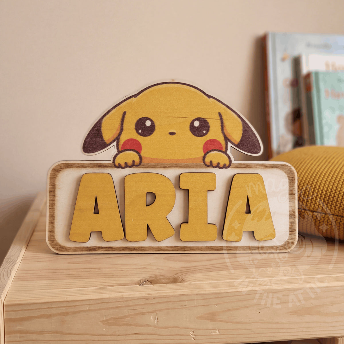 Placa de madera personalizada con Pikachu asomando por encima del nombre Aria en letras amarillas en relieve, apoyada en una estantería de madera junto a un cojín mostaza